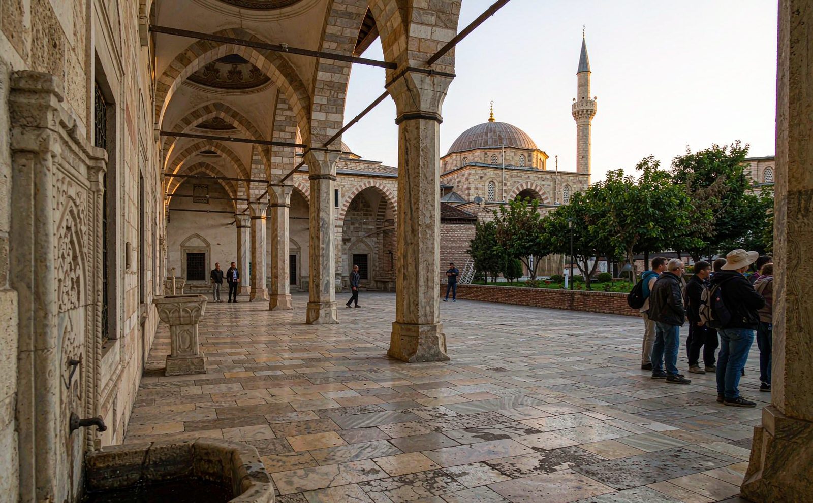 Visitors in an Ottoman mosque courtyard in Sarajevo, stone arcades and a slender minaret in morning light